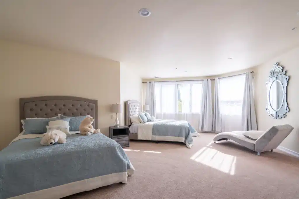 Two beds with blue bed covers with light shining through the window. There are 2 teddy bears sitting on a bed. There is a mirror on the wall and a recliner couch next to the window